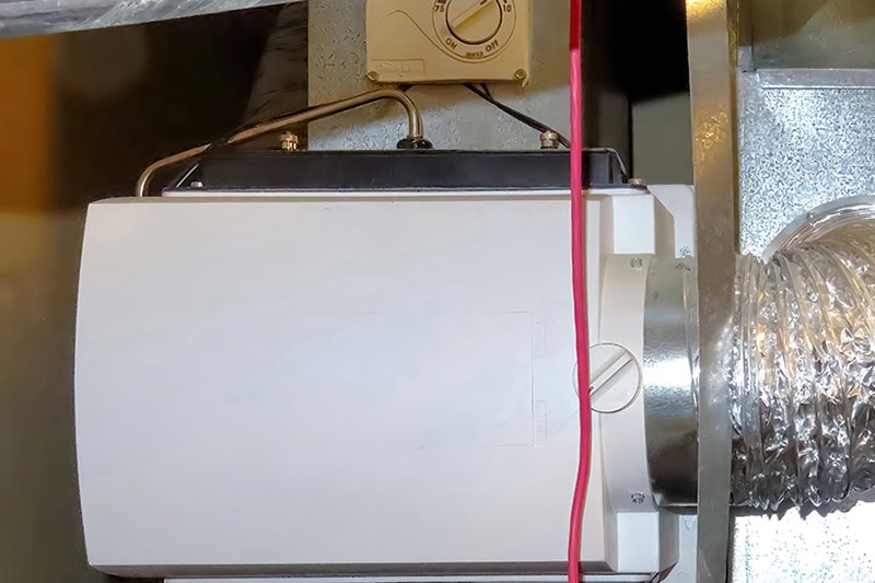 A close-up view of a whole-house bypass humidifier mounted on metal HVAC ductwork, featuring a white cover unit attached to flexible foil ducting on the right and a humidistat dial installed above it.