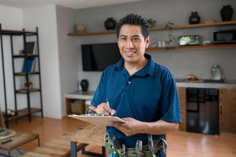 What Is a Plumbing Inspection? Electrician working at a house and looking at the camera smiling while writing on a clipboard - home improvement concepts